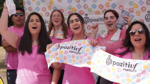 A group of people wearing pink shirts holding Pozitivf Fertility banners and items, standing in front of a Pozitivf Fertility backdrop with colorful shapes.