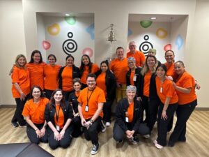 A group of people wearing orange shirts are posing for a photo in a room with light-colored walls and wooden flooring. The background features colorful abstract shapes on the wall and a bell mounted on a stand.